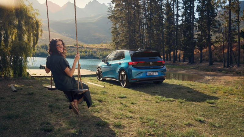 Una chica en un columpio junto a un Volkswagen ID.3 en un bosque con un lago al fondo