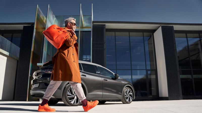 Una hombre con un bolso y abrigo naranja caminando delante de un Volkswagen ID.