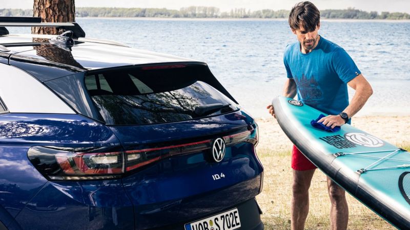 Un hombre con una tabla de surf en un lago junto a un Volkswagen ID.4 azul