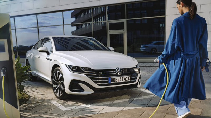 Chica de azul con un cargador frente a un Volkswagen Arteon híbrido eléctrico