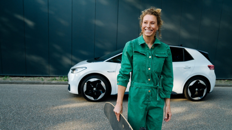 Chica co mono verde sonriendo de pie delante de un Volkswagen ID.3 blanco visto de costado