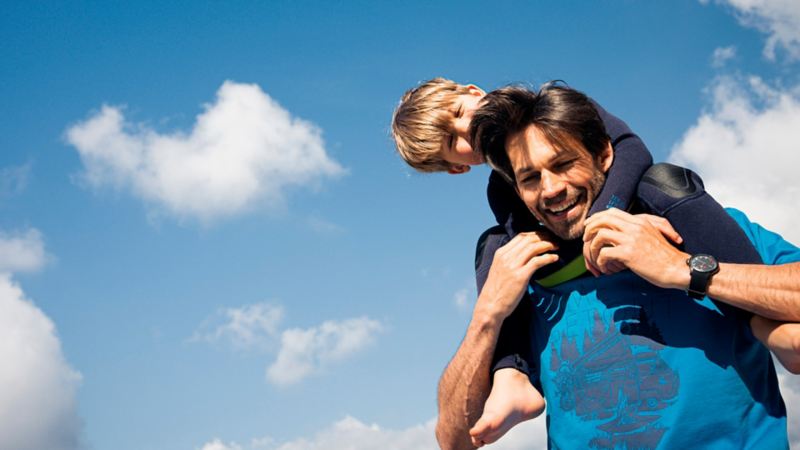 Un padre con su hijo en los hombros y un fondo de cielo azul con nubes