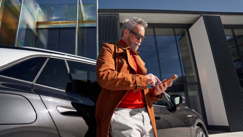 Un hombre con un móvil apoyado en un Volkswagen ID. frente a una fachada de cristal
