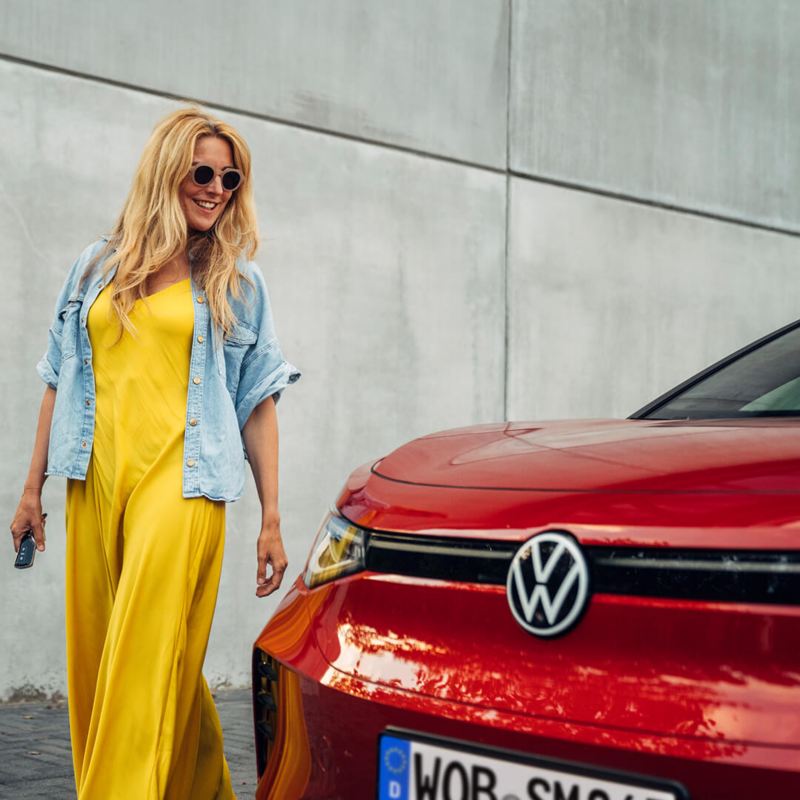 Chica de amarillo junto a un Volkswagen ID.4 rojo visto parcialmente frente a un muro de concreto