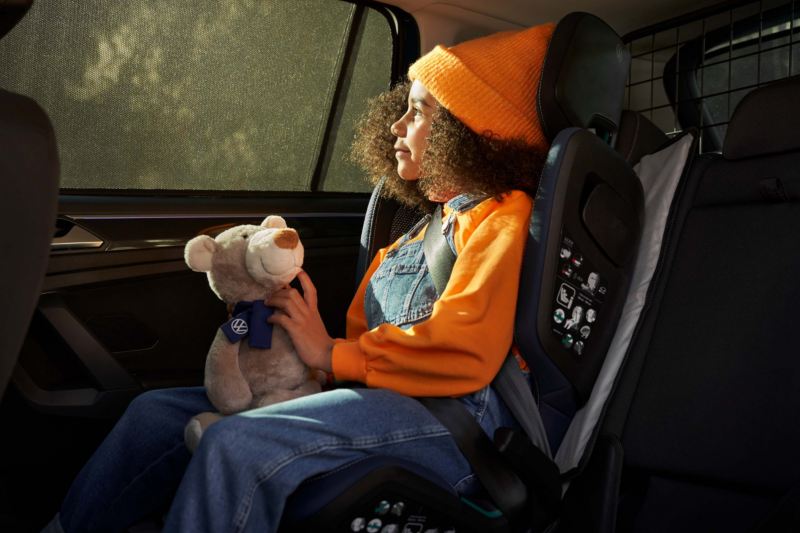 Una niña de naranja con un peluche en un asiento de niños en un Volkswagen