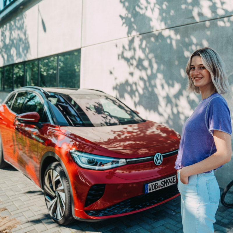 Chica sonriendo de pie frente a un Volkswagen ID.4 rojo bajo la sombra de un árbol