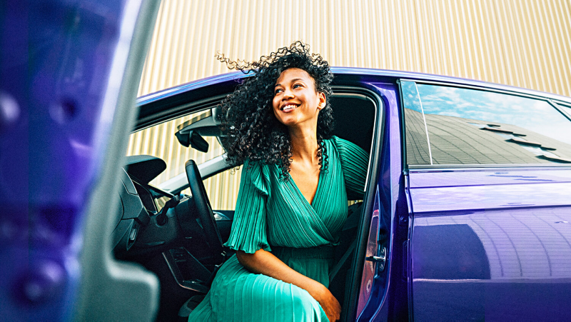 Chica de verde sonriendo sentada en el puesto del conductor de un Volkswagen azul con la puerta abierta