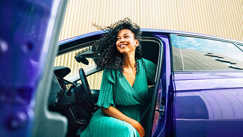 Chica de verde sonriendo sentada en el puesto del conductor de un Volkswagen azul con la puerta abierta