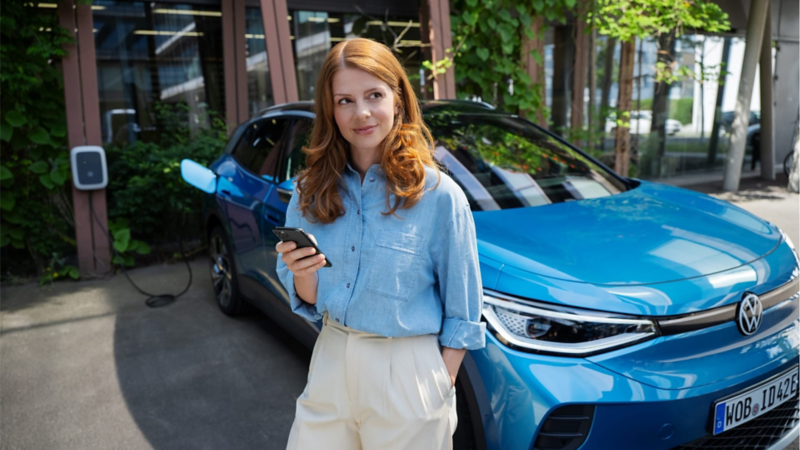 Una mujer con un móvil de pie frente a un Volkswagen SUV eléctrico de color azul