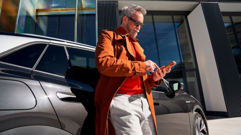 Un hombre con abrigo naranja mirando el móvil apoyado en un Volkswagen color gris