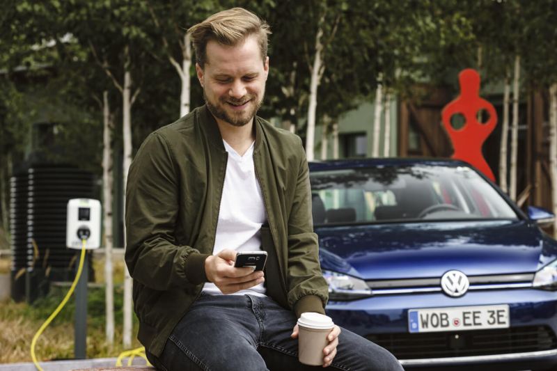 Un hombre con un móvil y un café esperando que se cargue su coche eléctrico