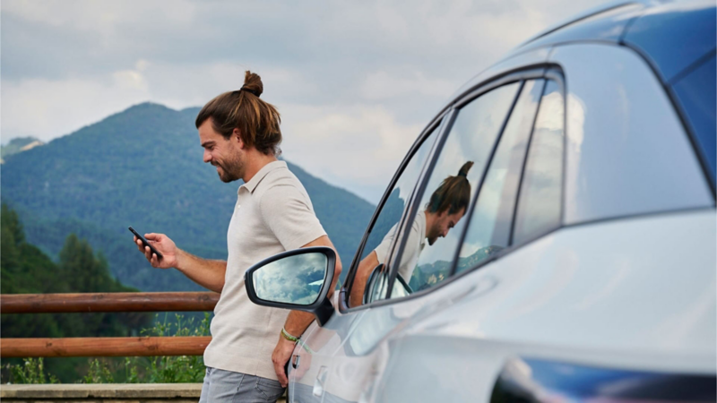 Un chico con un móvil apoyado en un Volkswagen SUV eléctrico en la montaña