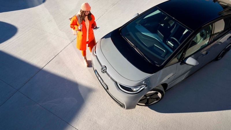 Chica de naranja viendo el móvil junto a un Volkswagen ID.3 gris visto desde arriba