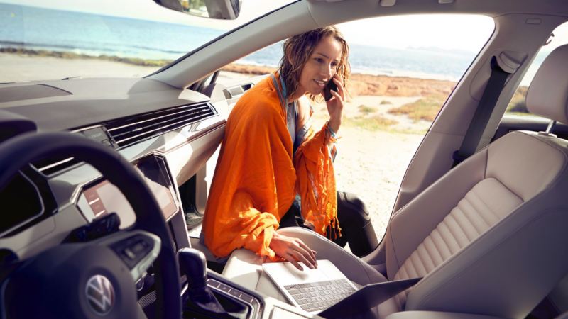 Chica hablando por móvil y con ordenador sentada en el borde de la puerta abierta de un Volkswagen