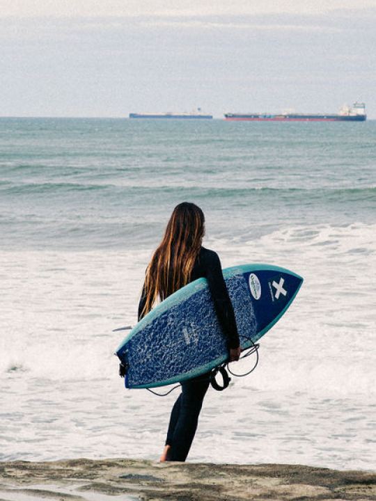 Una chica con una tabla caminando hacia el mar con el The Roadcycled Wetsuit de Blue Banana