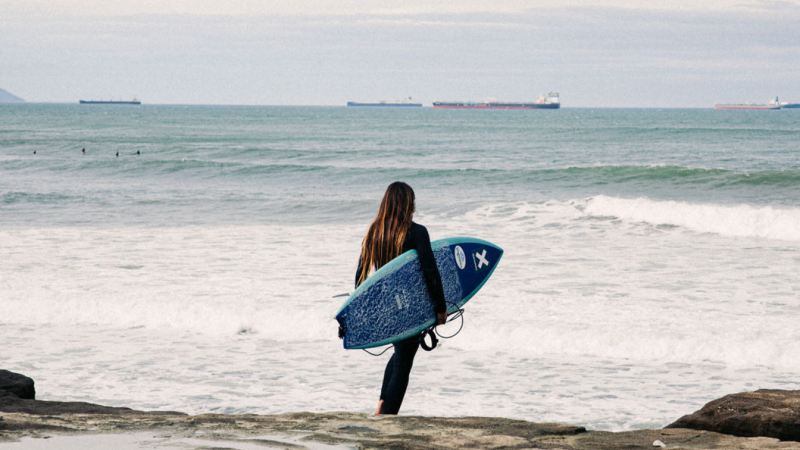 Una chica con una tabla caminando hacia el mar con el The Roadcycled Wetsuit de Blue Banana