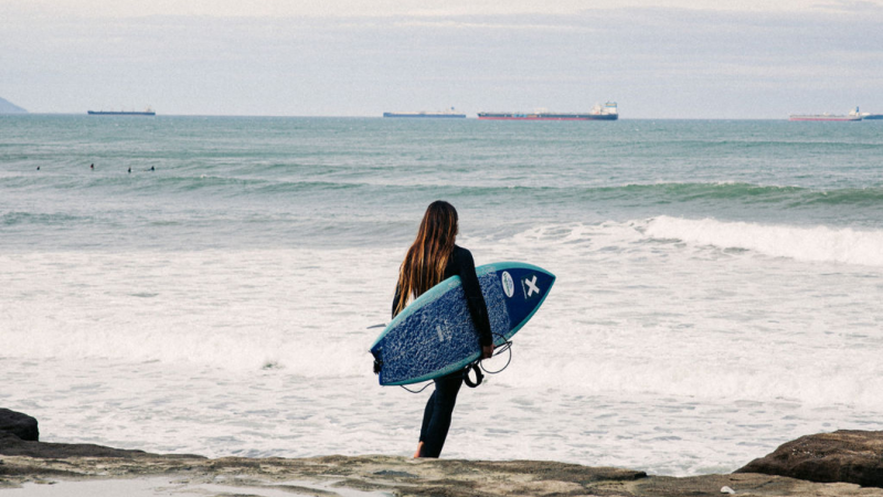 Una chica con una tabla caminando hacia el mar con el The Roadcycled Wetsuit de Blue Banana