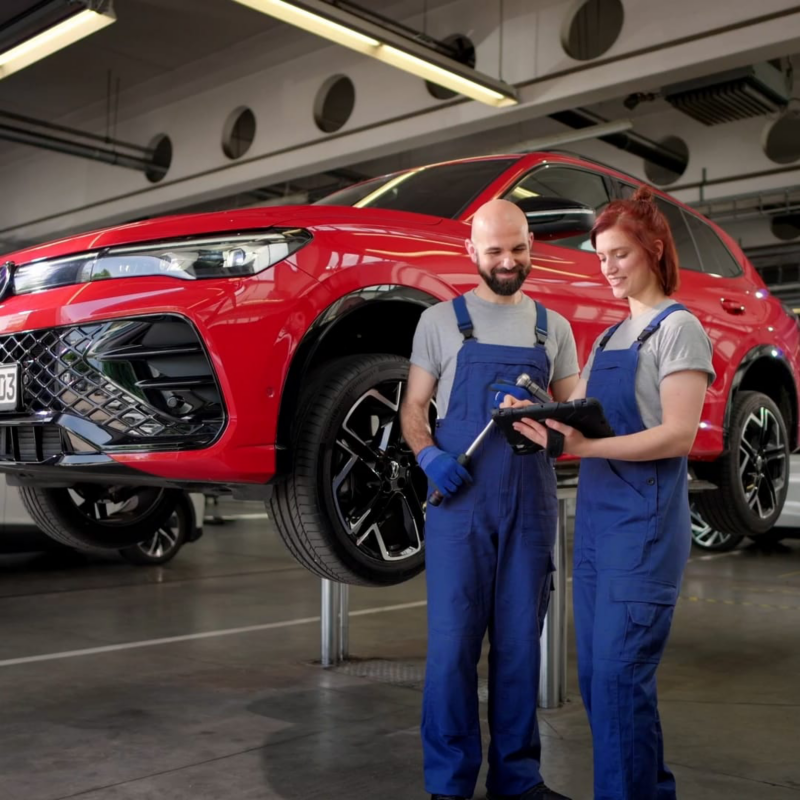 Dos mecánicos charlando en el taller con un Volkswagen rojo elevado detrás