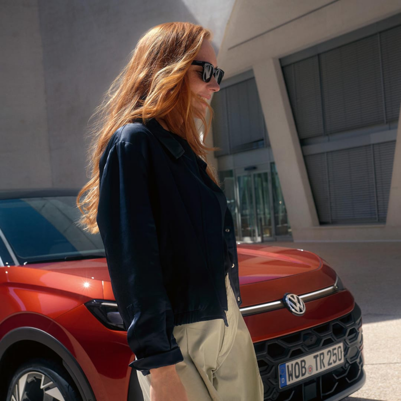 Una chica sonriente con gafas de sol pasando por delante de un Volkswagen rojo