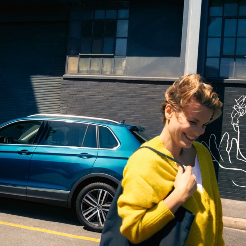Mujer de amarillo sonriendo delante de un Volkswagen azul aparcado en la calle