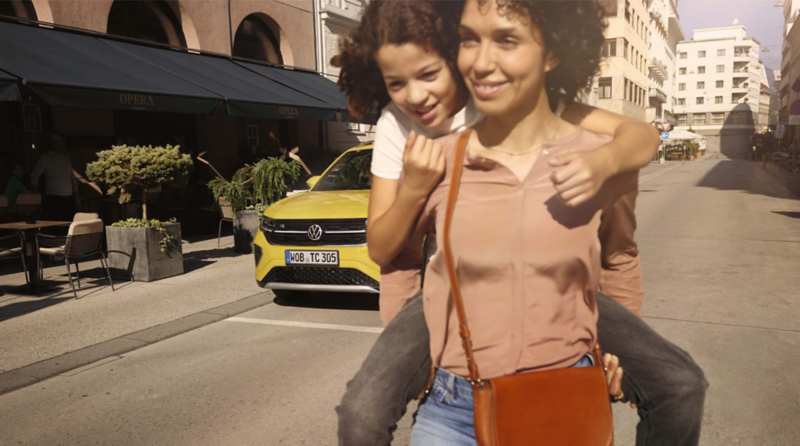 Una mujer cargando una niña en su espalda con un Volkswagen amarillo detrás