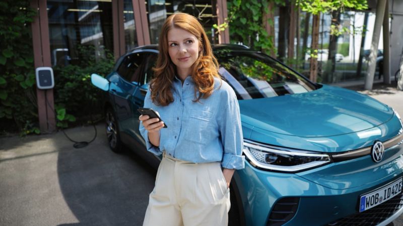 Una chica usando un móvil delante de un Volkswagen ID.4 azul conectado a un cargador