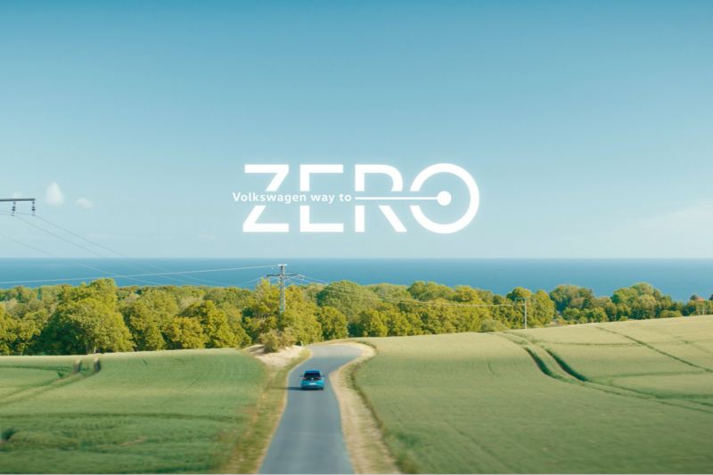 Un Volkswagen visto de frente circulando por una carretera en el campo y el mar de fondo con el logo de Way to Zero sobre impreso