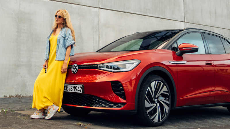 Chica de amarillo apoyada en un Volkswagen ID.5 rojo frente aun muro de concreto 