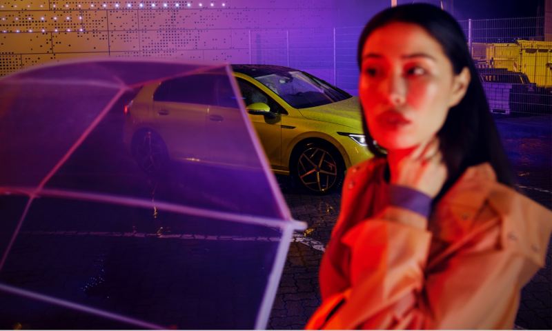 Chica desenfocada delante de un Volkswagen Golf 8 amarillo aparcado de noche
