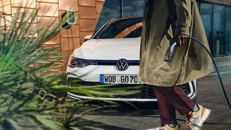 Vista inferior de un chico caminando delante de un Volkswagen Golf 8 GTE blanco con un cable de carga en la mano