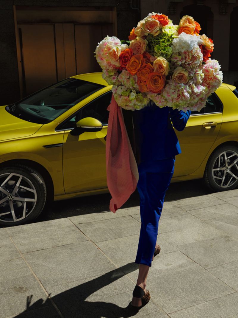 Chica caminando con un gran ramo de flores hacia un Volkswagen Golf amarillo aparcado en la calle