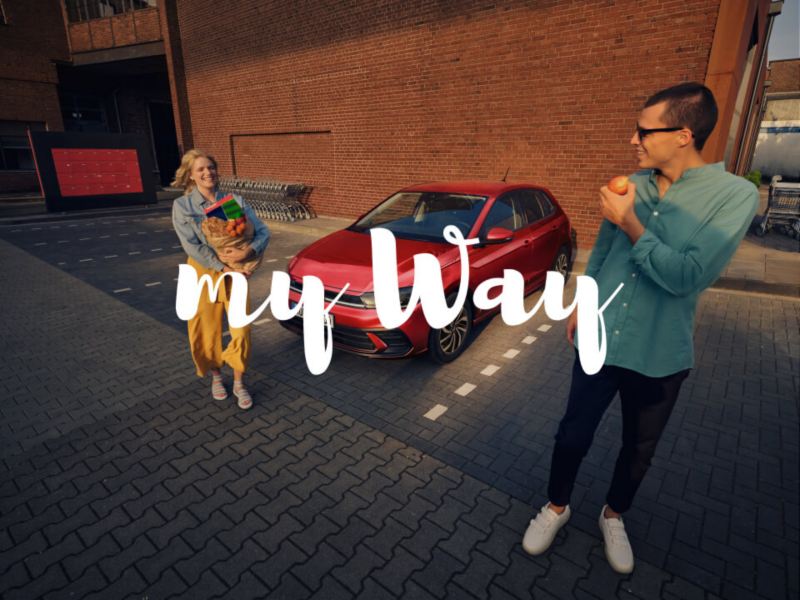 Un chico y una chica delante de un Volkswagen de color rojo estacionado con el logo de My Way