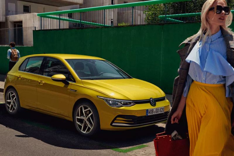 Una chica delante de un Volkswagen Golf 8 amarillo aparcado en la calle