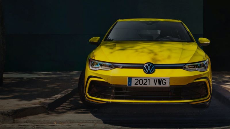 Vista del frontal de un Volkswagen Golf 8 amarillo aparcado a la sombra de un árbol