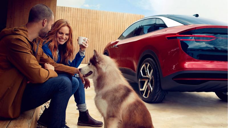 Un Volkswagen ID. Crozz rojo en posición diagonal desde atrás junto a un chico, una chica con una taza de café y un perro.