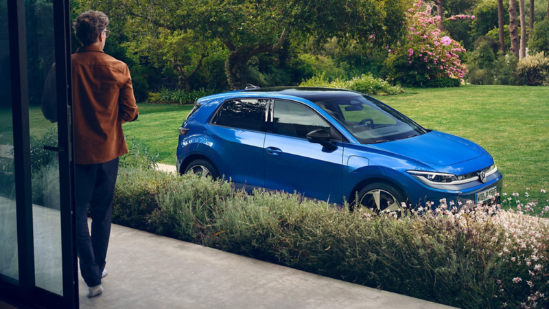 Vista a través de una puerta de cristal abierta hacia un jardín verde; un Volkswagen ID. Polo azul está estacionado en la entrada.