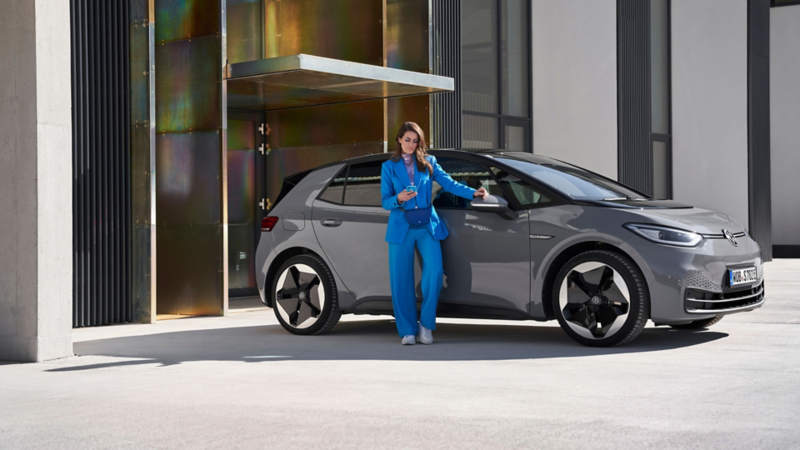 Una chica de azul  con un móvil en la mano de pie delante de un VW ID.3 gris