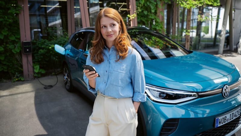 Una chica con un móvil delante de un Volkswagen ID.4 azul conectado a un punto de carga