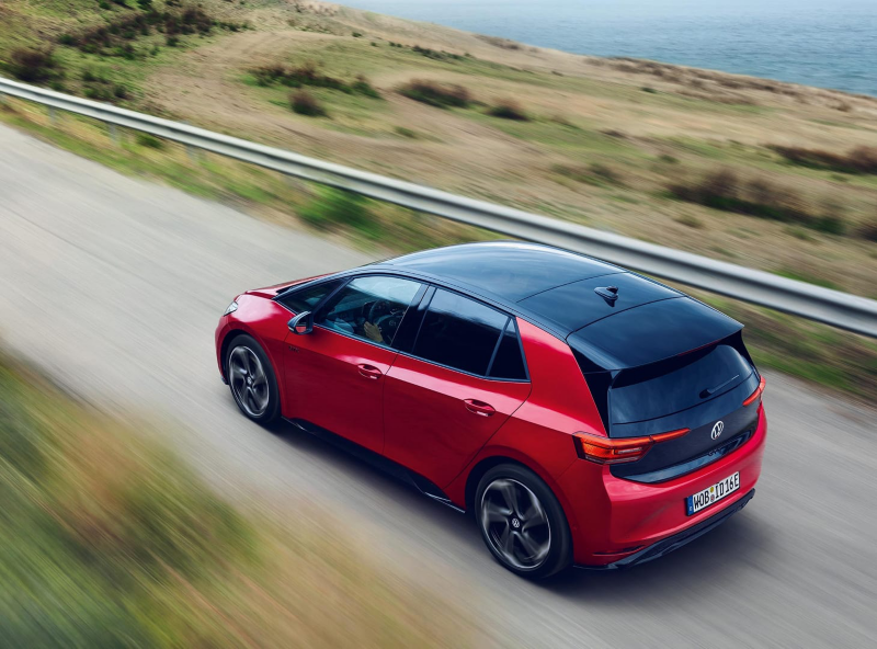 Plano lateral posterior de un ID.3 rojo en camino por una carretera con un paisaje desenfocado y el mar