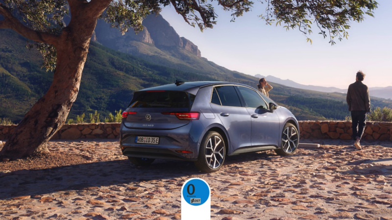 El nuevo ID.3 Neo de Volkswagen aparcado en un exterior natural al sol mientras dos personas disfrutan de la vista, con la etiqueta Cero Emisiones