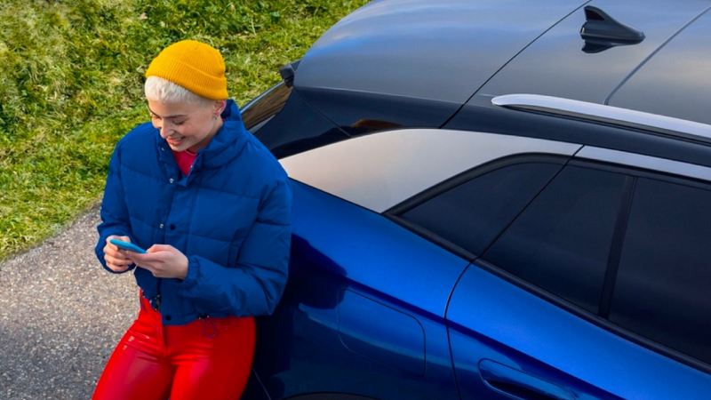 Chica mirando el móvil apoyada en la parte trasera de un Volkswagen ID.4 azul