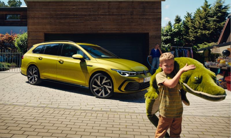 Un niño con un peluche delante de un Volkswagen Golf 8 Variant amarillo aparcado en casa