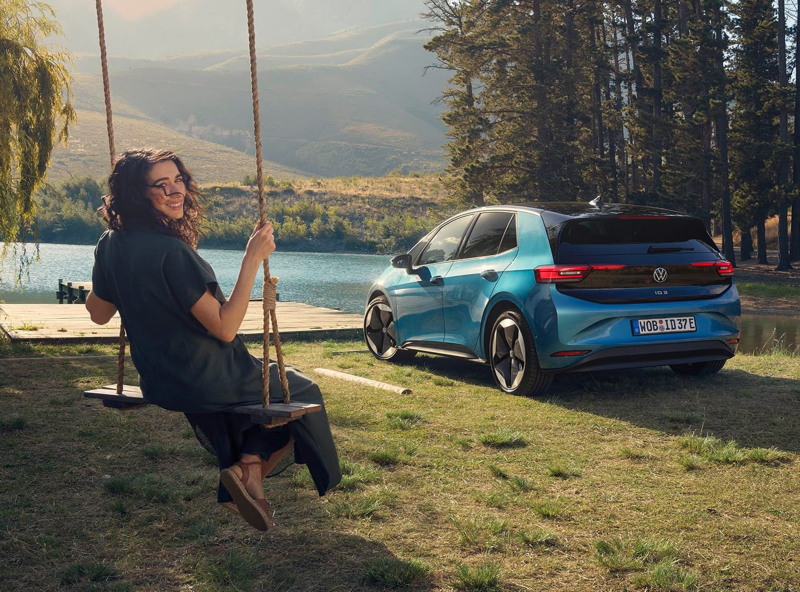 Una chica subida en un columpio al lado de un Volkswagen eléctrico de color azul estacionado al lado de un lago