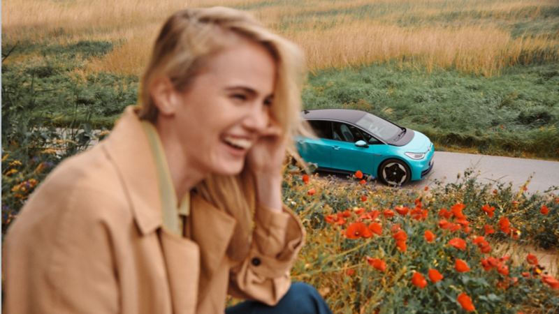 Chica en primer plano sonriendo en un campo de flores delante de un Volkswagen ID.3