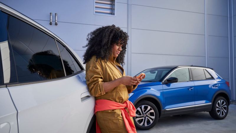 Una chica usando el móvil apoyada en un Volkswagen blanco y un T-Roc azul de fondo
