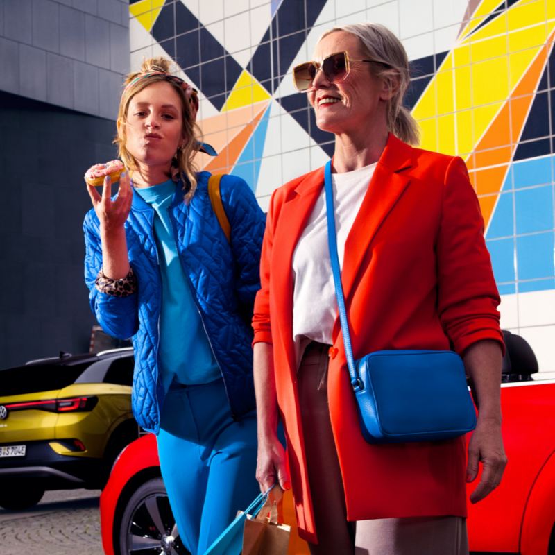 Dos chicas caminando delante de dos coches y un muro de colores