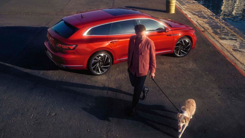 Un hombre con un perro delante de un Volkswagen Arteon Shooting Brake rojo