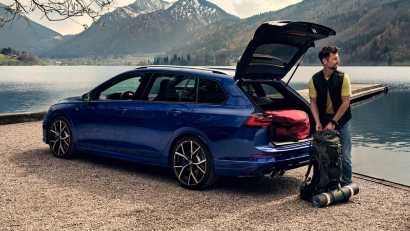 Un hombre junto a Volkswagen Golf 8 Variant R con el maletero abierto junto a un lago con montañas de fondo