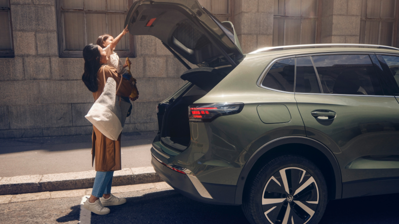 Una niña con su madre cerrando el maletero de un Volkswagen de color verde estacionado en la calle