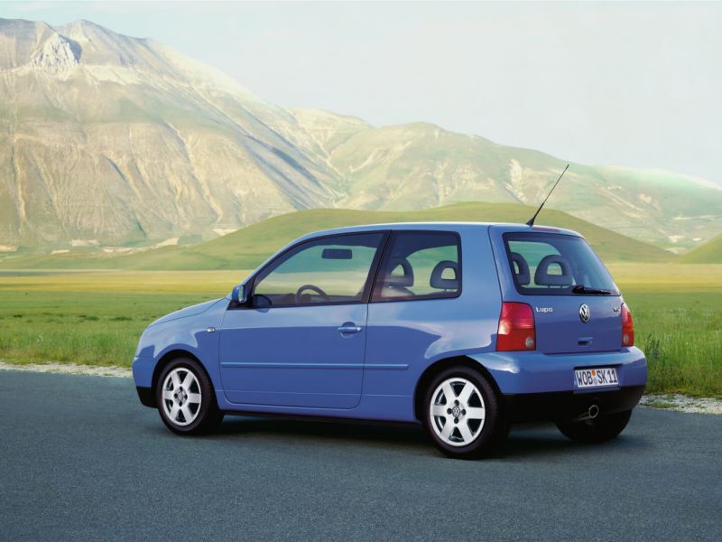 Volkswagen Lupo azul visto de costado en una carretera frente a las montañas